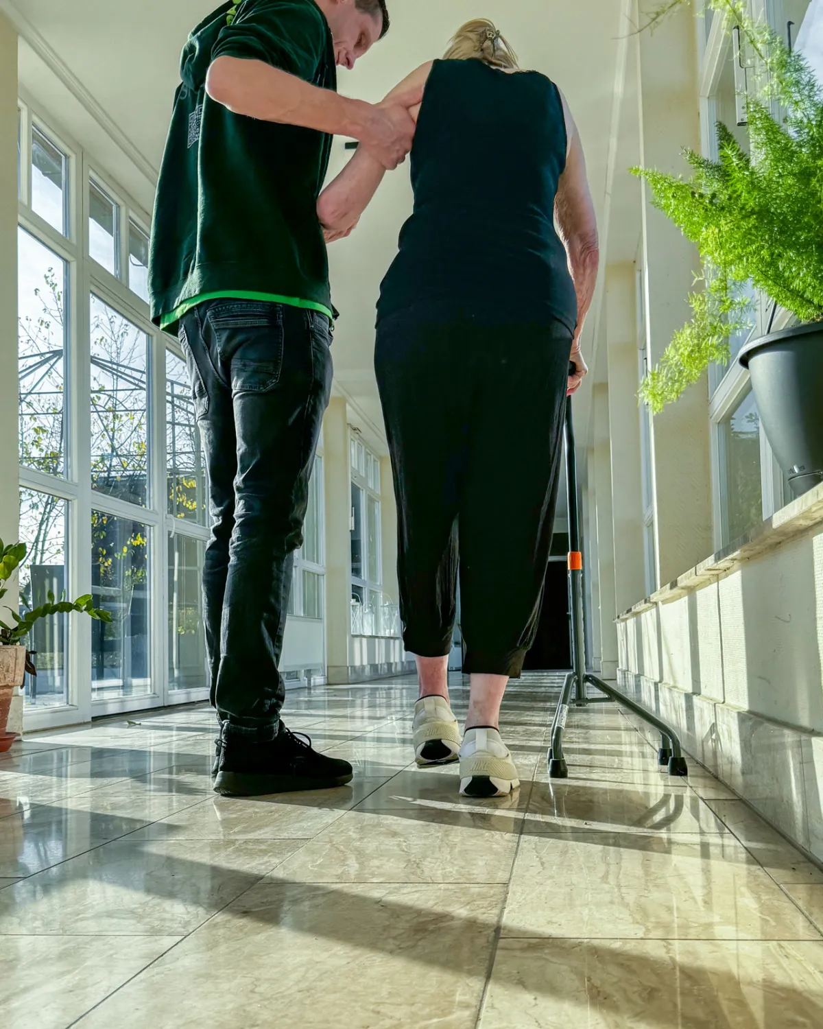 Ein Therapeut geht mit einer Bewohnerin am Rollator durch einen hellen Flur