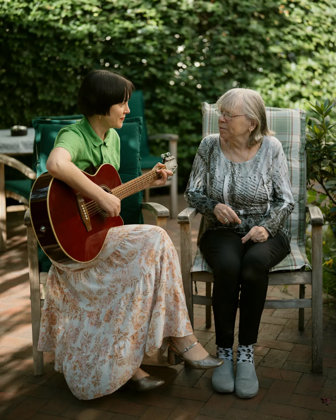 Therapeutin spielt auf der Gartenterrasse einer Einrichtung Gitarre, eine Bewohnerin sitzt neben ihr und hört zu