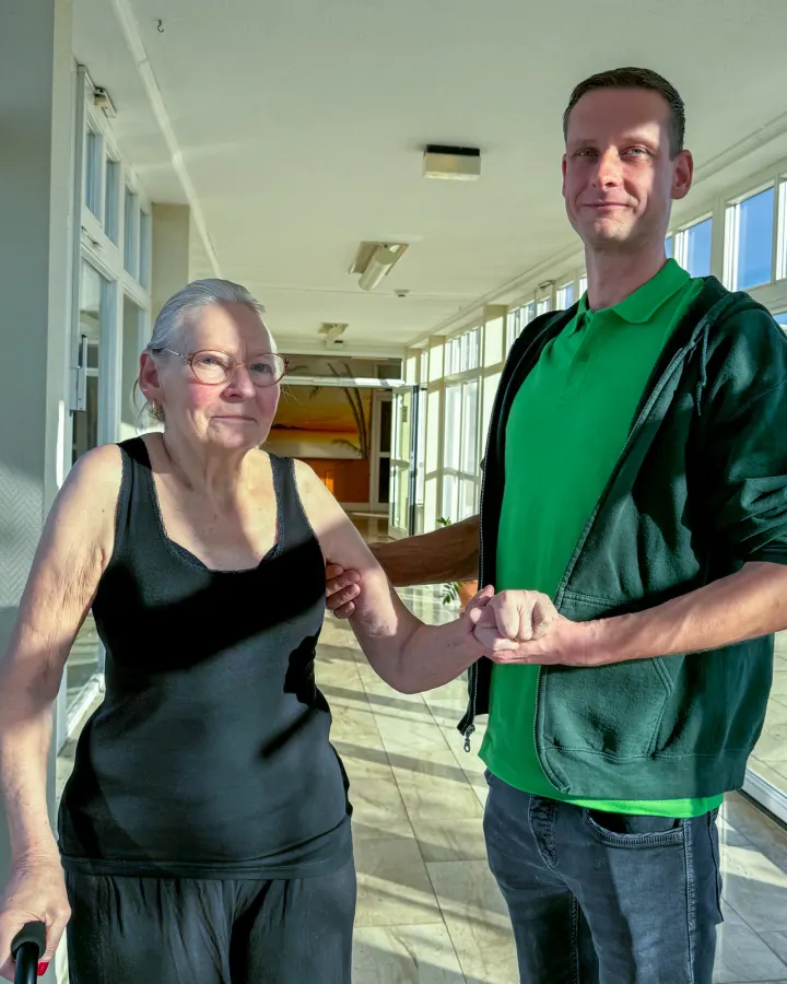 Therapeut und Bewohnerin gehen Hand in Hand einen hellen Flur entlang