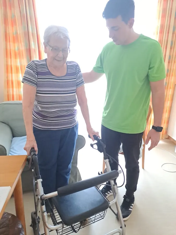 Ein Therapeut im grünen Shirt begleitet eine Bewohnerin am Rollator